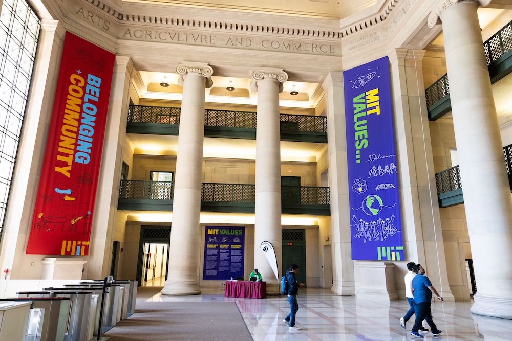 Three people walk through Lobby 7. There is a tall, red banner that says Belonging and Community, with illustrations of the Dome, a person playing piano, and arms hugging a heart. A purple banner says MIT Values... and has illustrations of a smiling beaver, a globe, and people wearing graduation caps who have their arms in the air and appear to be jumping.