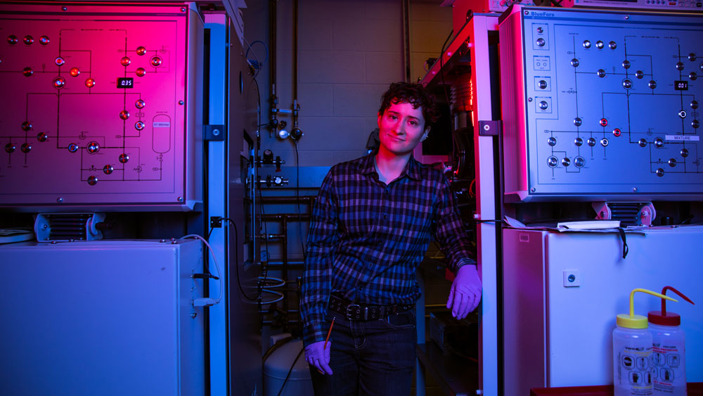 With dramatic pink and blue lighting, Alex Greene stands in between two large machines with patchbays on front, inside a lab.