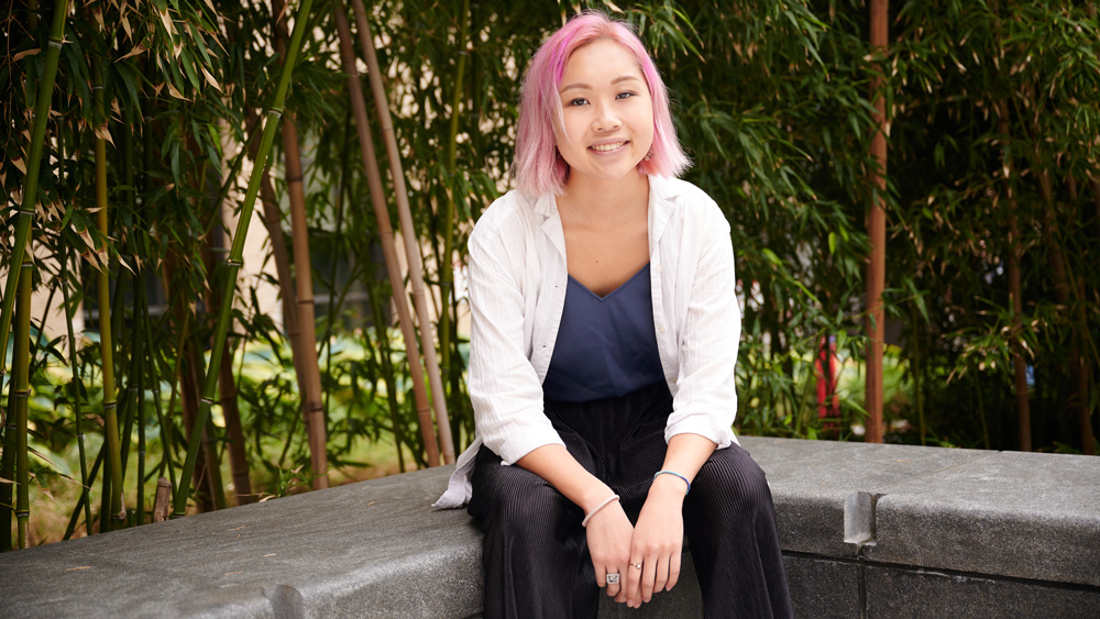 Susan Su sits on a concrete bench with tall bamboo in background and wears a Brass Rat ring.