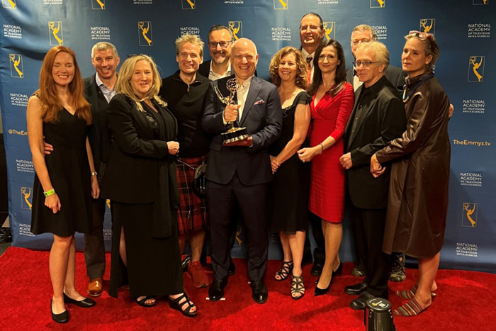 12 people smile for a photo, with a man in center holding the Emmy Award