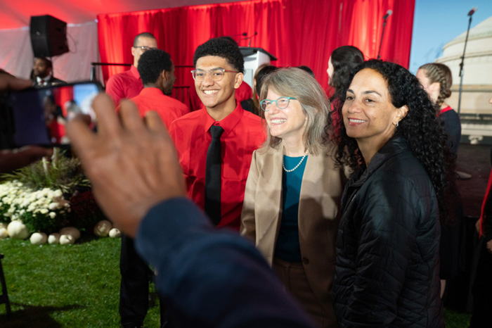 President-elect Sally Kornbluth poses for a photo with 2 people. Peeking out from the left side, are the hands and camera screen of the photographer taking a photo.