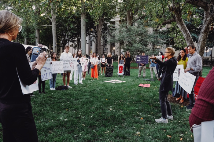 Photo of Linda Griffith with a bullhorn, addressing a semicircle of about 30 rally attendees on a grassy area, many holding hand-painted signs.
