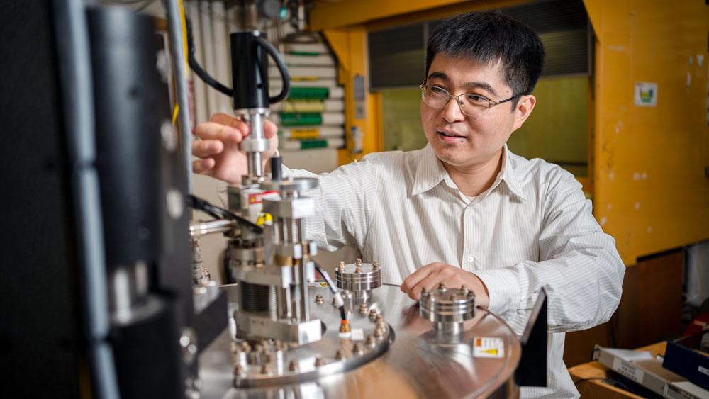 Luqiao Liu adjusts a part of a large cylindrical device, the sputtering deposition system.