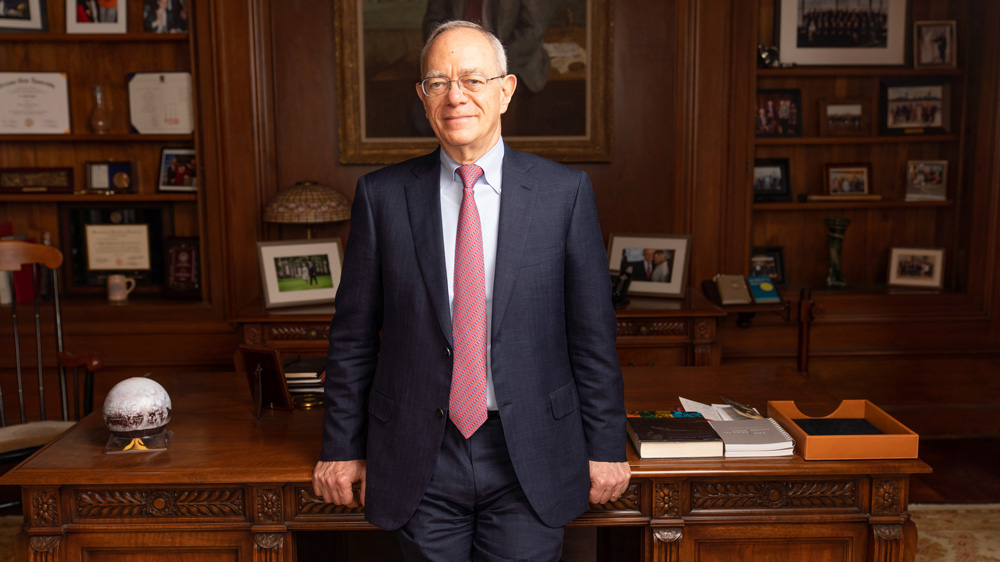 L. Rafael Reif in President’s office, with hands resting on large wooden table. The wooden walls and shelves are filled with mementos and photos.
