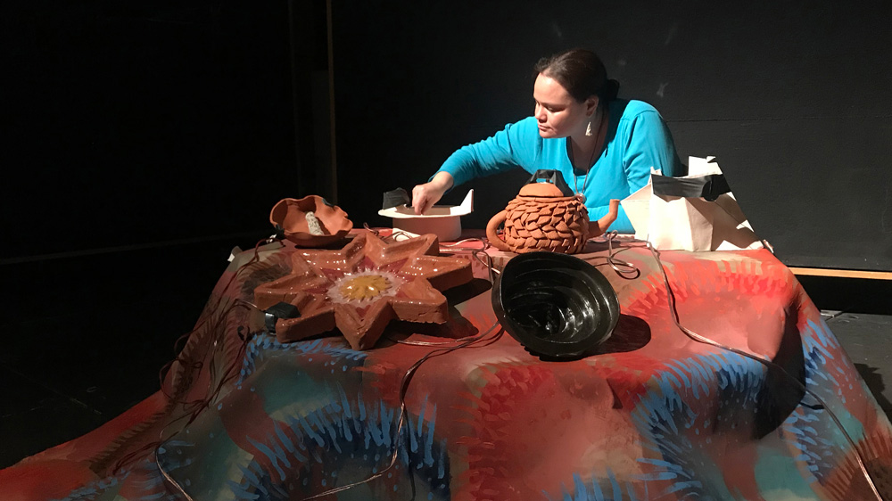  Erin Genia arranges 5 pieces of pottery on a fabric stand, under a spotlight