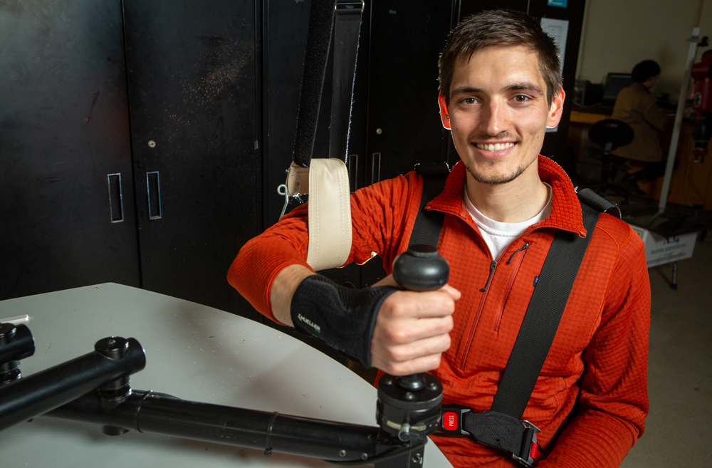 Hermus looks at camera while his arm is in a sling used for testing. He grips a joystick while wearing a black wrist strap.