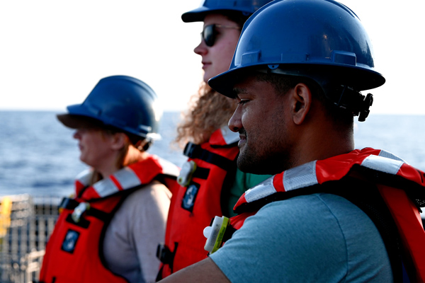 3 people on boat wearing orange life jackets and chatting, with ocean in background