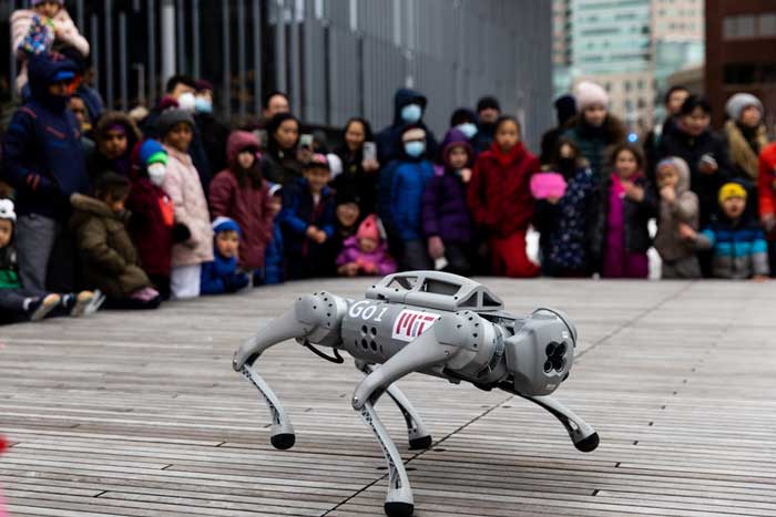 A large crowd watches as a robot dog demo. Its grey and has a big MIT logo and has 4 sensors where a head would be.