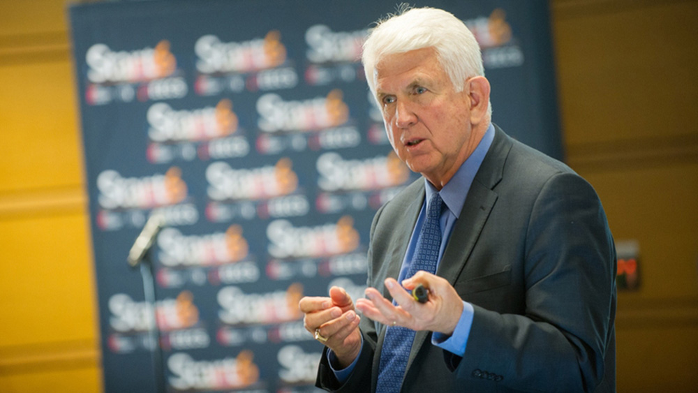 Bob Metcalfe speaks at an event, with blurry banner in background