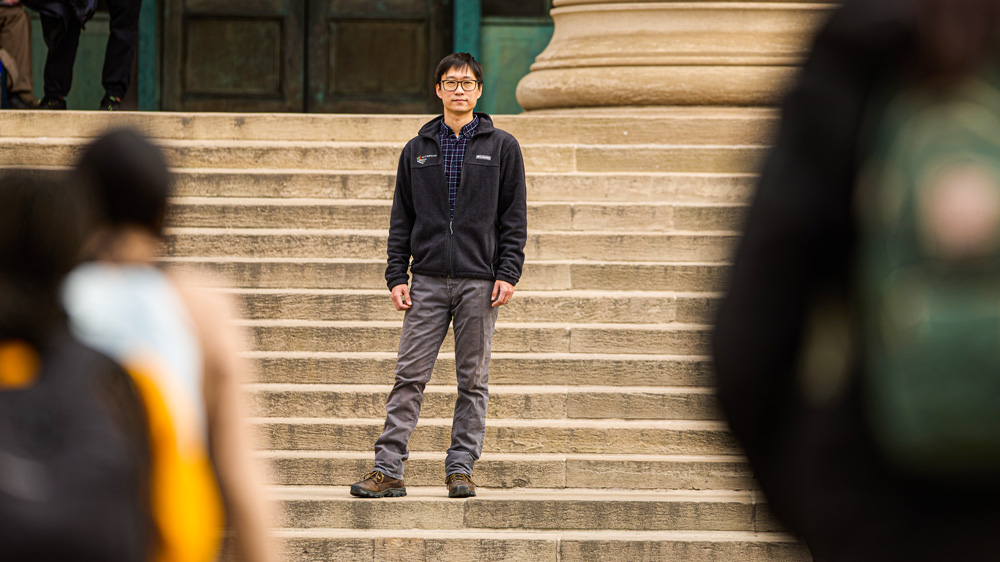 Guo stands on the steps of 77 Mass. Ave, and in the foreground are the blurred people framing the image.