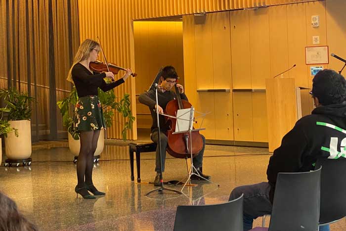 A violinist and cellist perform in a lobby inside an MIT building, with glimpses of seated audience