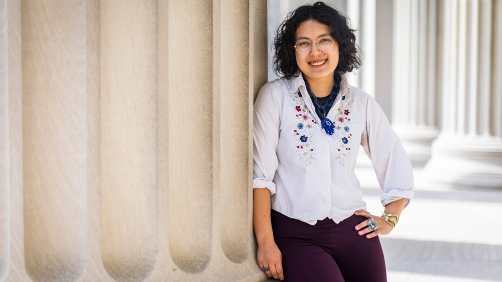 Dogan leans against a beige column while on MIT campus, and wears a Brass Rat ring. More columns are in the background.