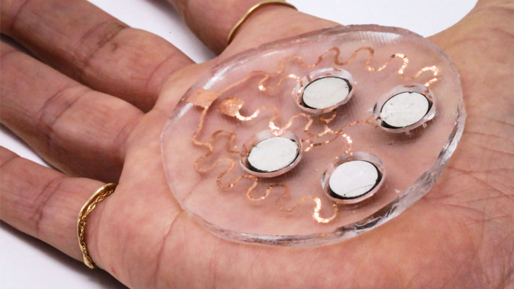 On the palm of a person’s hand is a unique circular device. The base looks like clear gel pad with 4 circular white sensors on it. Wavey copper lines attach the sensors to each other.