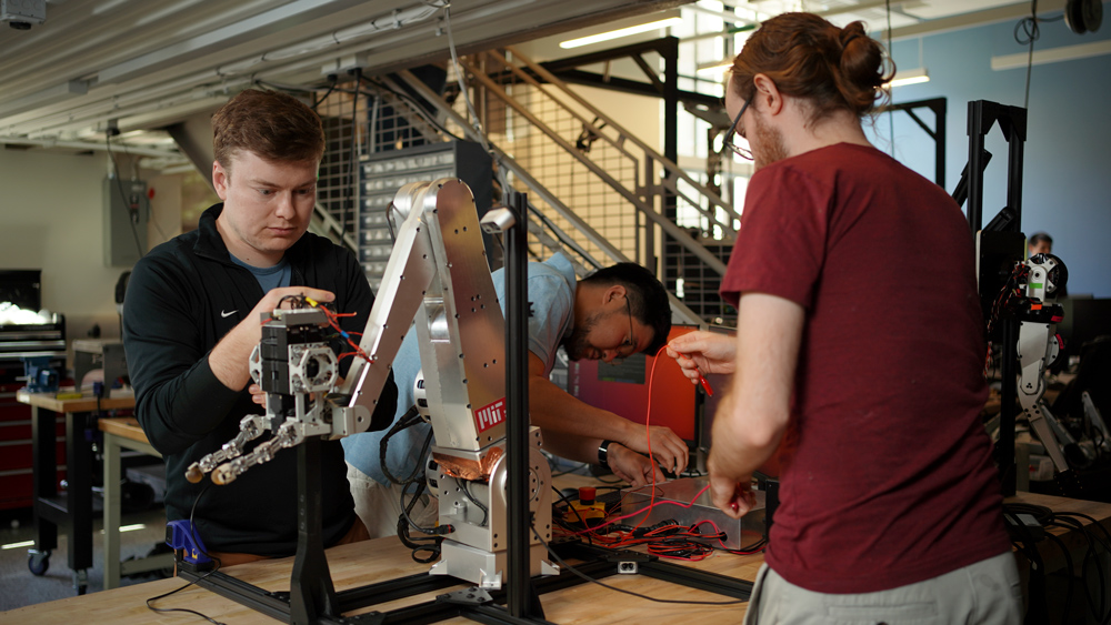 Andrew SaLoutos, Hongmin Kim, and Elijah-Stanger-Jones working on a robot arm in a lab