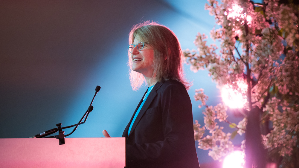 Sally Kornbluth speaking at a podium with a pink flowering tree behind her