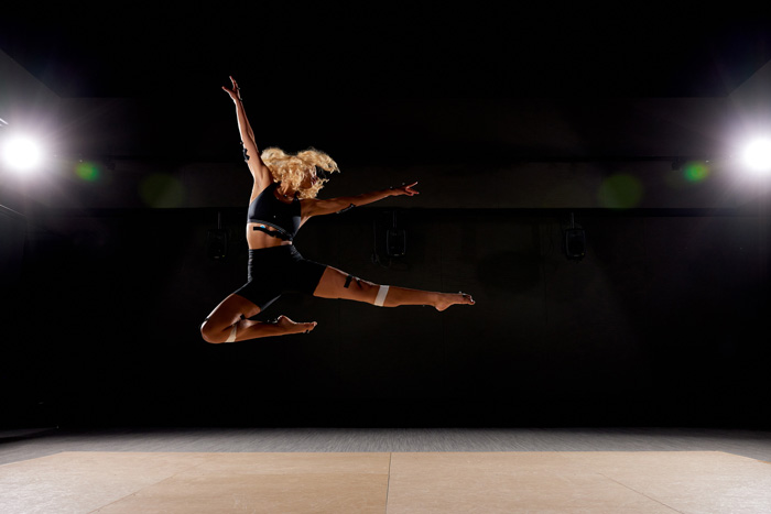 Against a dark background with spotlights on the left and right, Kaelyn Dunnell jumps in a dynamic pose. Sensors are attached to Dunnell with black tape.