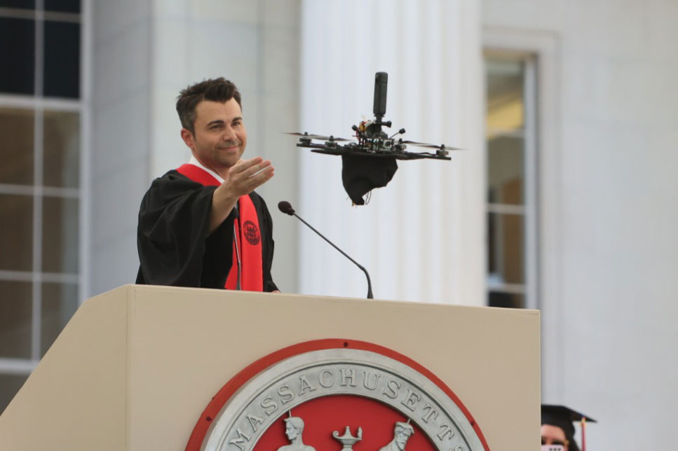 Mark Rober gestures to a drone, which is flying up with his graduation cap