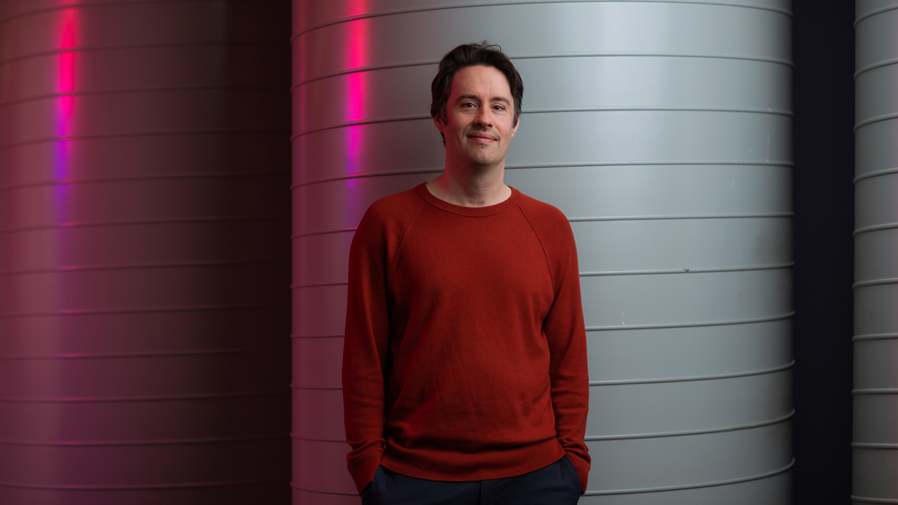 Harlow portrait as he stands in front of giant cylindrical white pipes. A strong pink light shines from the left. 