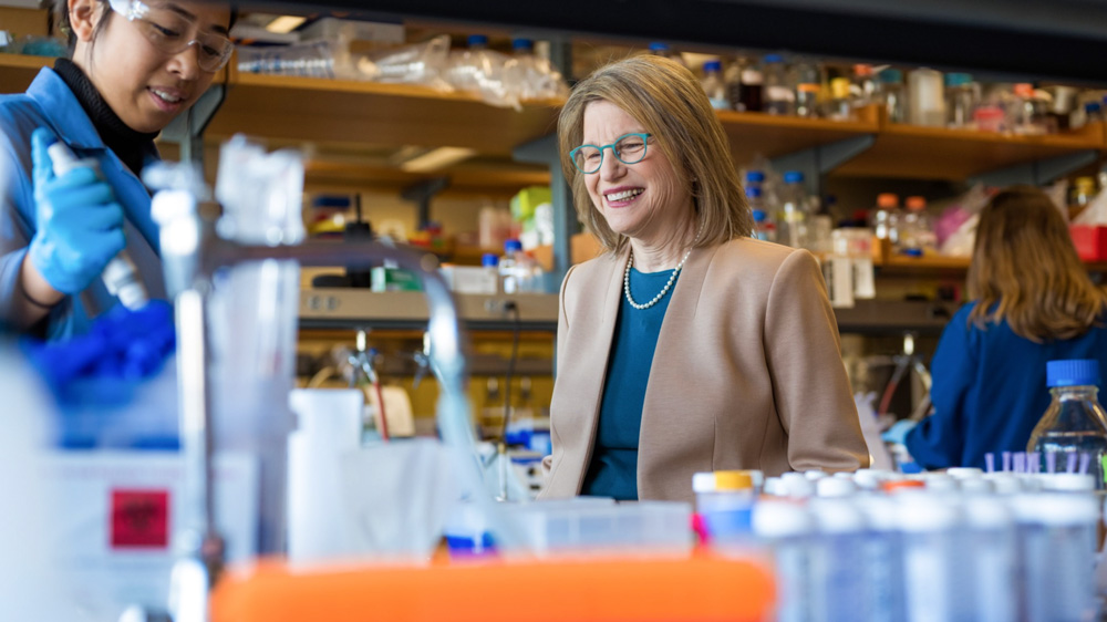 Sally Kornbluth, center, visits a lab where scientists are working, and watches as a scientist in blue protective coat and gloves uses a pipette