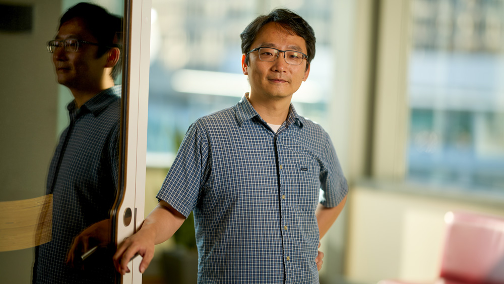 Andy Sun poses for portrait during the daytime next to reflective door while resting their right hand on handle. The blurry background has glass window with view of a building, a red lounge chair, and a round table. 