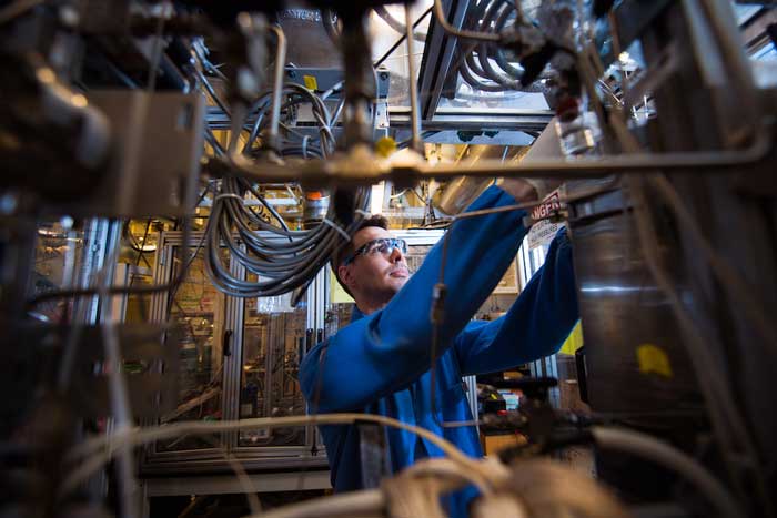 Jamison Watson, wearing safety glasses, adjusts the trickle-bed reactor, comprising a variety of pipes and wires in the foreground of the image.
