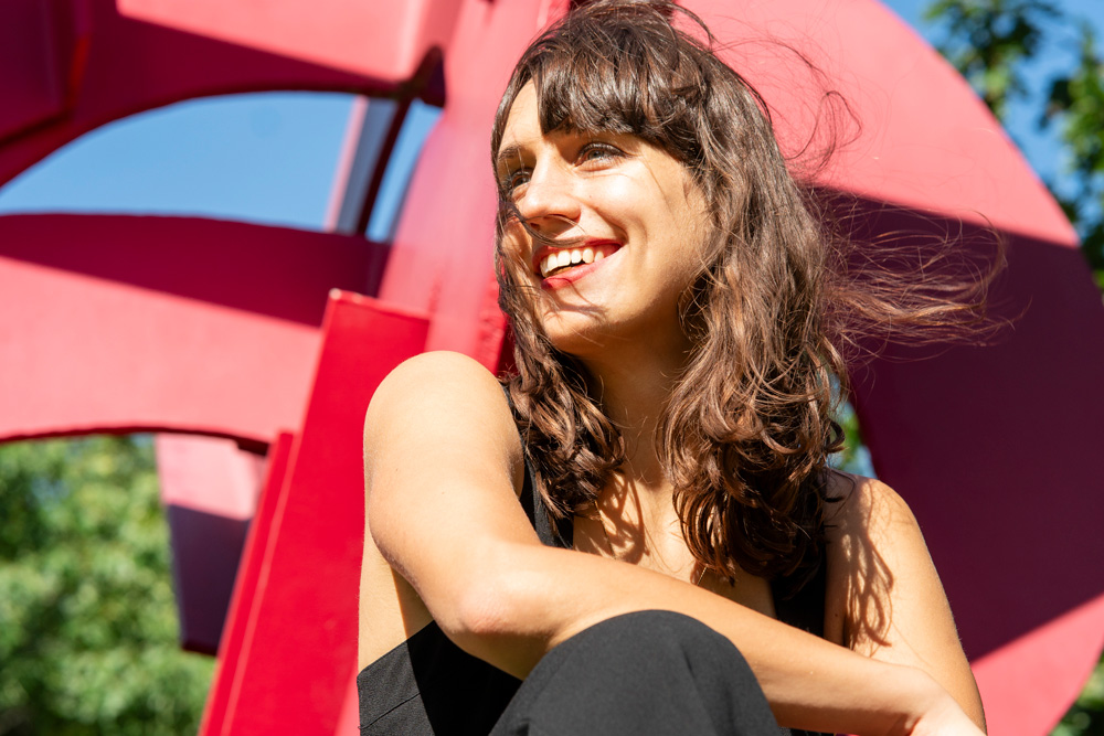 Jaye smiles while crouching, and a red sculpture,, Mark di Suvero’s Aesop’s Fables, II, 2005, is in the background