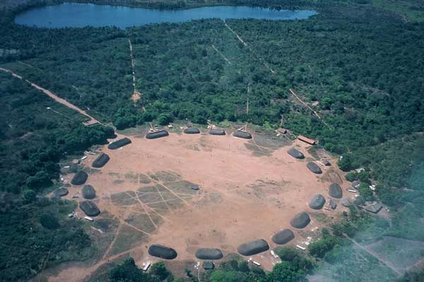 The village has about 20 huts that form a large a ring around an empty, brown, circular area. Lots of trees are around the village.