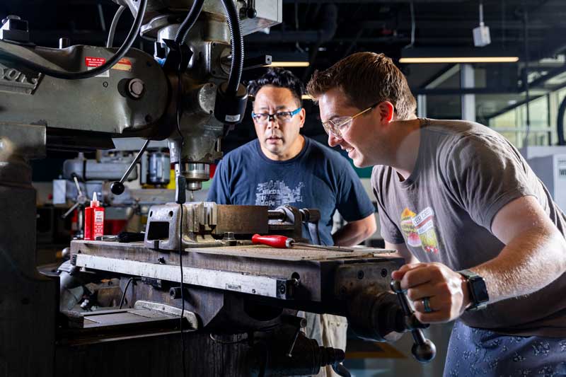 Hayami Arakawa and Randall Briggs use large machine and stare at a drill-bit inside the Hobby Shop.
