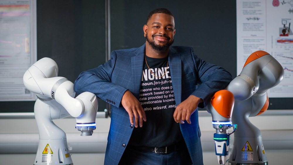 Michael West smiles while in between two robot arms. His elbows rest on the two machines, and his black shirt has the definition of “Engineer.”