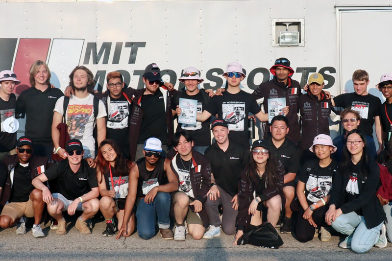 About 25 Motorsports team members smile, with an "MIT Motorsports" logo on wall behind them