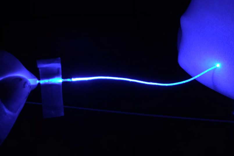 A blue glowing fiber in darkness. The fiber is held by fingers and seems to light up with it touches another hand.