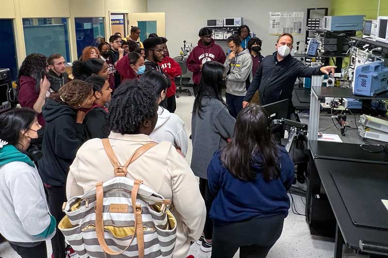 While about 30 students look on, an instructor points to a machine in a lab filled with oscilloscopes and other equipment. 