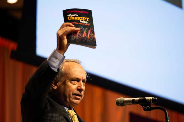 Rodney Brooks holds a book about ChatGPT in the air while at a podium