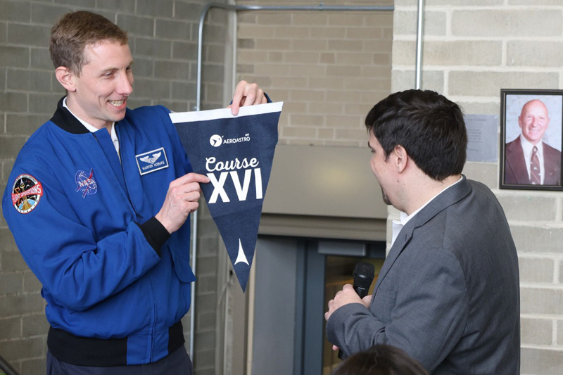Woody Hoburg, left, wears a NASA jacket and holds a triangular flag that says "MIT AeroAstro Course XVI" and shows it to another person