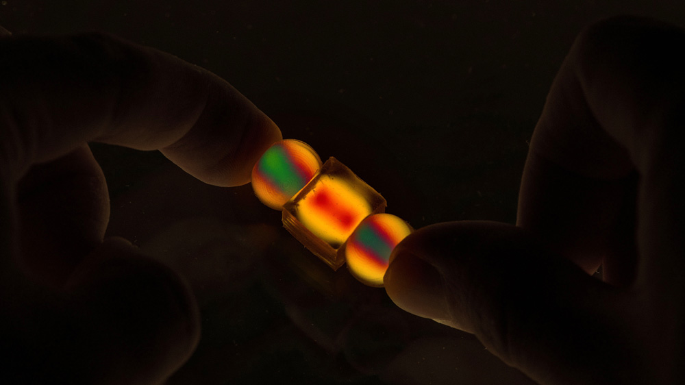 In darkness, thumbs hold 2 small spherical and 1 small cube objects that light up in vivid yellow, red, and green.