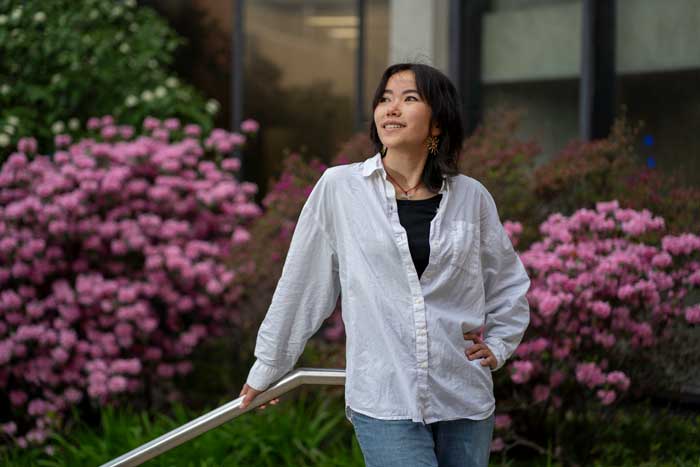 Cindy Xie smiles outside and looks up and to the side. The background has blooming pink rhododendrons.