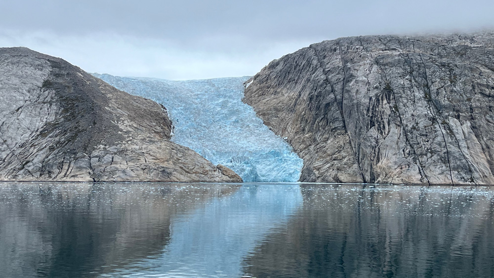 A blue glacier flows into a fjord between rocking hills