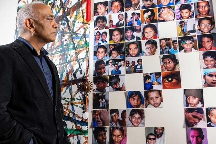 Pawan Sinha looks at a wall of about 50 square photos. The photos are pictures of children with vision loss who have been helped by Project Prakash.