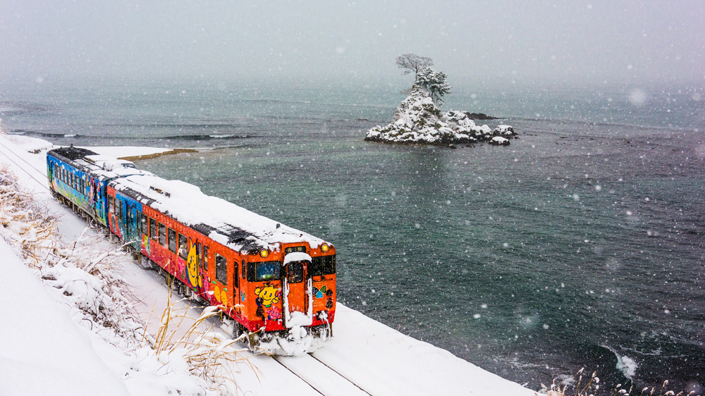 A colorful Japanese train moves through a snowy landscape near the ocean in Noto.