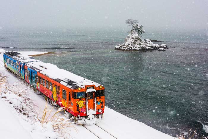 A colorful Japanese train moves through a snowy landscape near the ocean in Noto.