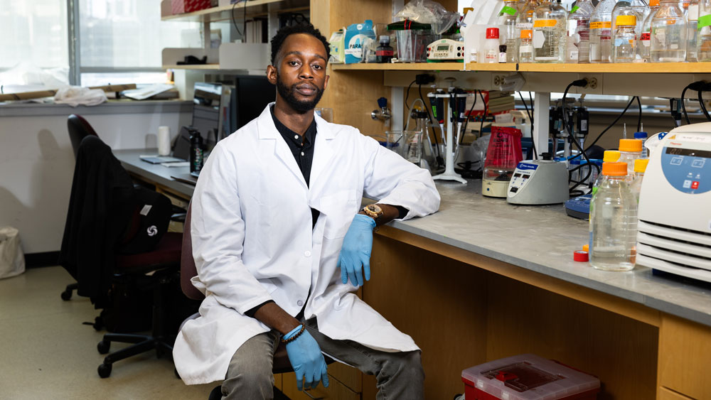 Feyisayo Eweje wears lab coat and gloves while sitting in a lab.