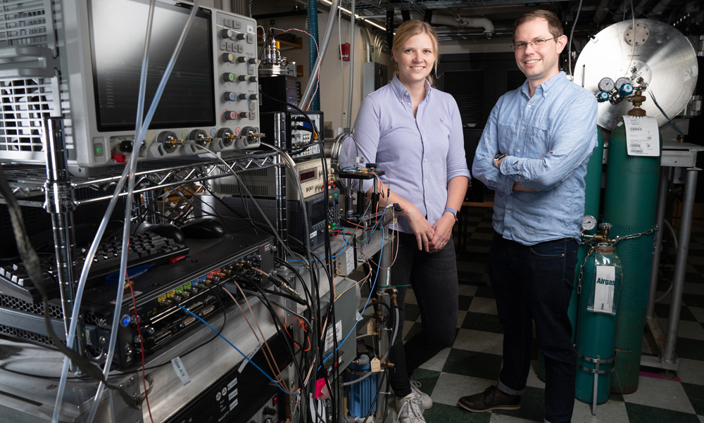Gabi Wenzel and Brett McGuire stand in their lab full of equipment on racks.