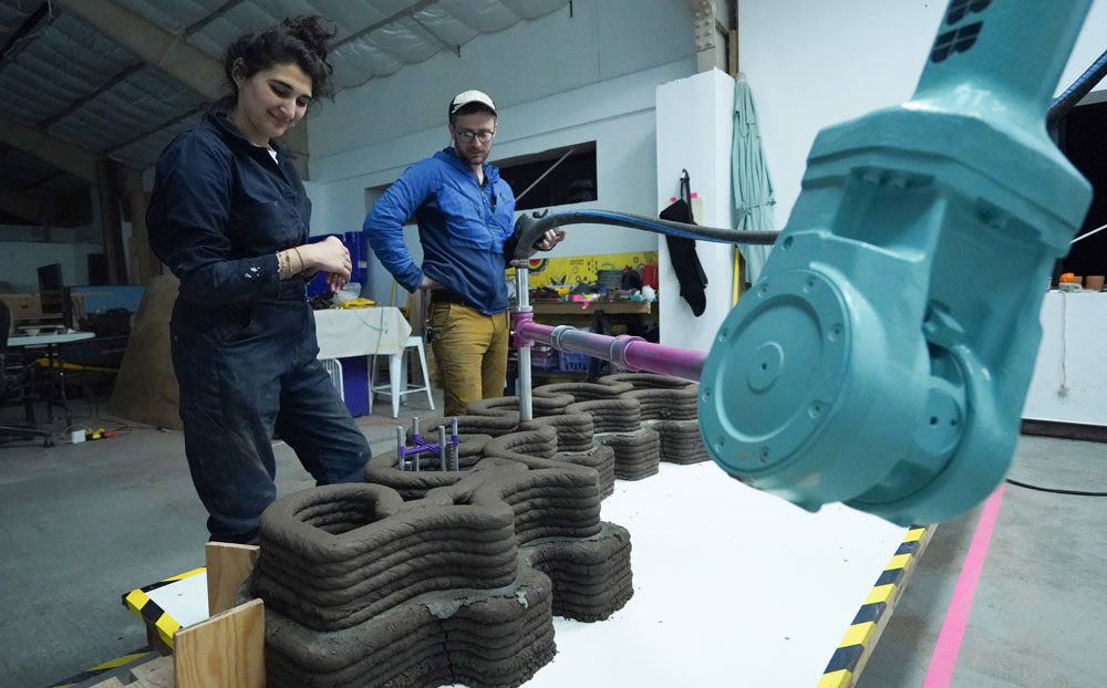 2 people watch a robot arm print a 3D cement wall.