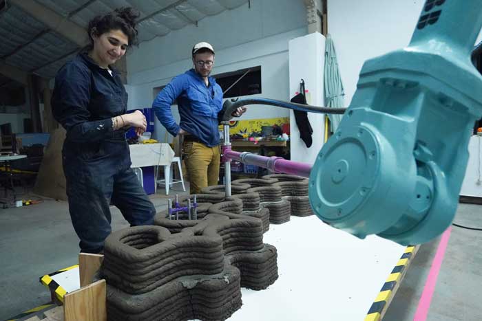 2 people watch a robot arm print a 3D cement wall.