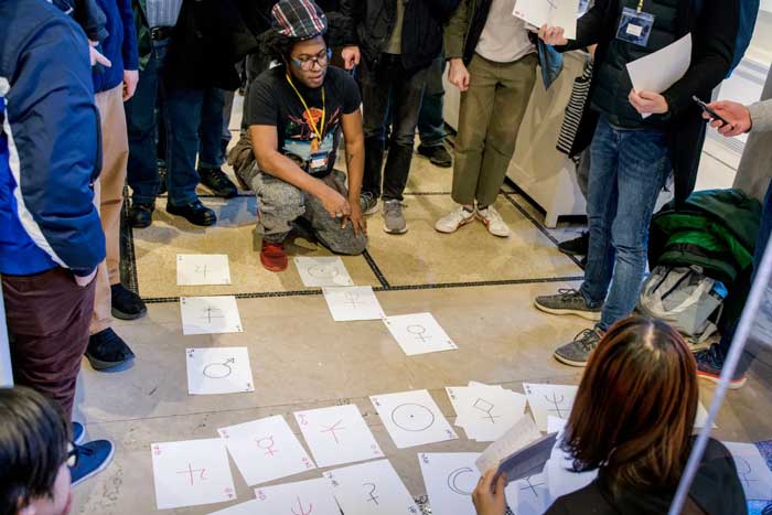 About 15 people gather round large papers on the floor with symbols on them