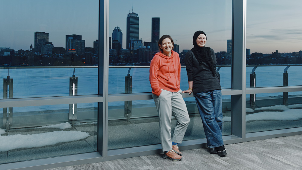 The two stand indoors by windows with Charles River in background