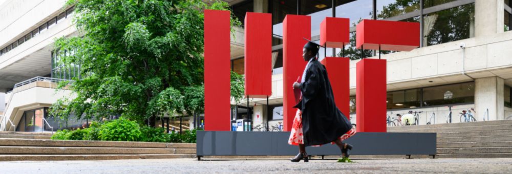 MIT student walking on graduation day in front of a large, red MIT logo.