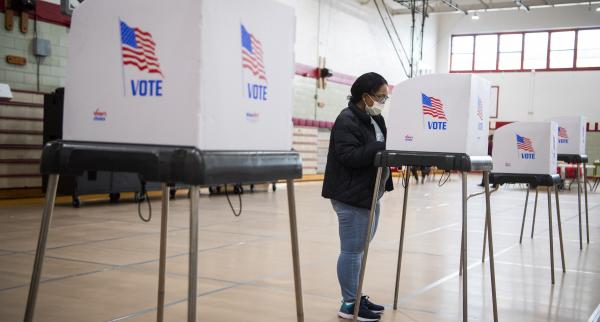 A woman stands at a voting booth inside a gym. The booth is white, with "VOTE" printed on it underneath the American flag.