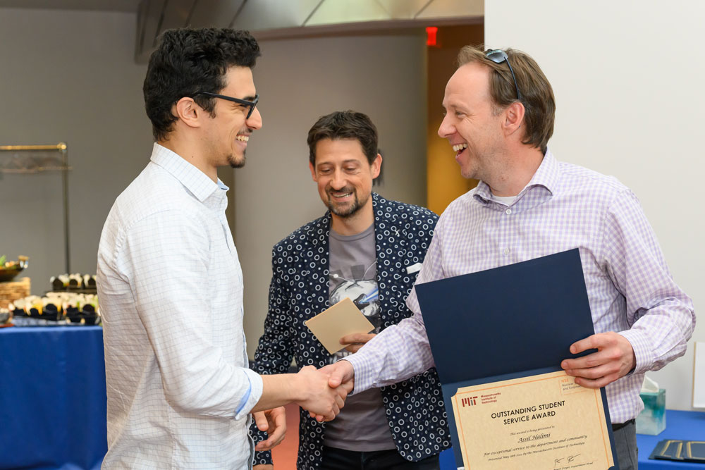two men shaking hands, the one of the right holding a certificate in a folder, third man between the two looking on, MIT