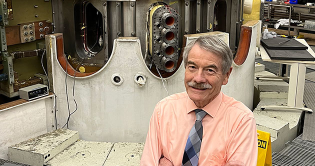 Male faculty member Ian Hutchinson in front of Alcator C-Mod, a fusion tokamak in a large lab sapce, MIT
